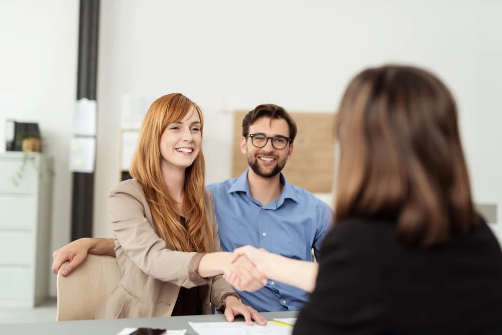 Happy young couple meeting with a broker in her office leaning over the desk to shake hands, view from behind the female agent-1